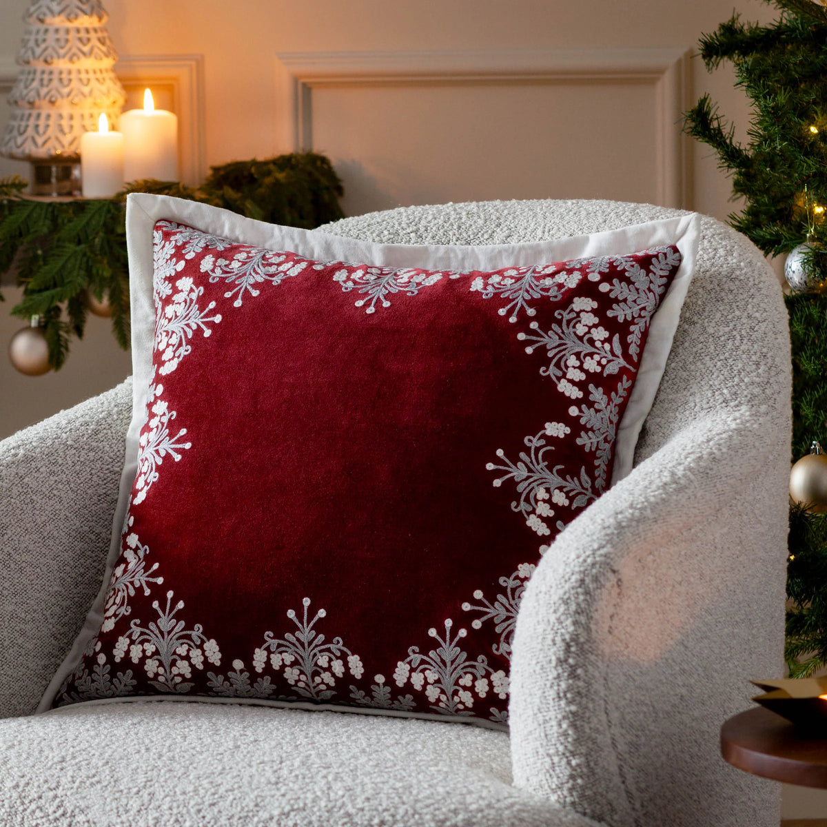 Red embroidered winter bloom cushion styled on a cream boucle armchair beside lit candles, a small Christmas tree with baubles and greenery, set in a warmly lit living room – Kumperi