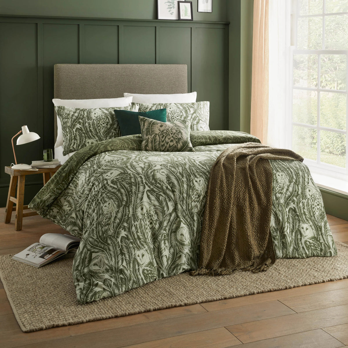 Forest green owl print duvet cover set styled on a bed with matching pillowcases, a plain dark green velvet cushion and a smaller owl print cushion, with a brown textured throw draped across the bed against a dark green panelled wall – Kumperi