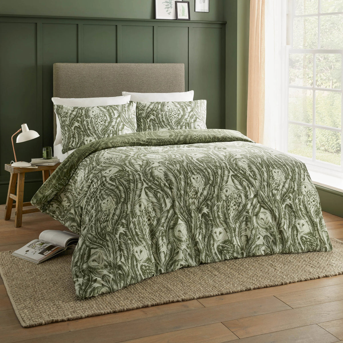 Front view of a bed dressed in the forest green owl print duvet cover set with matching pillowcases, styled against a dark green panelled wall with a wooden side table and lamp – Kumperi