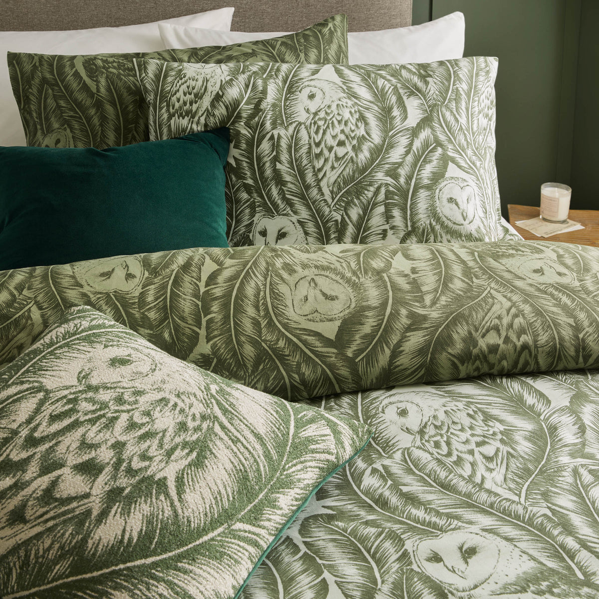 Close-up of stacked forest green owl print pillowcases and duvet folds showing the barn owl and banana leaf pattern in fine detail, with a dark teal velvet cushion beside them – Kumperi