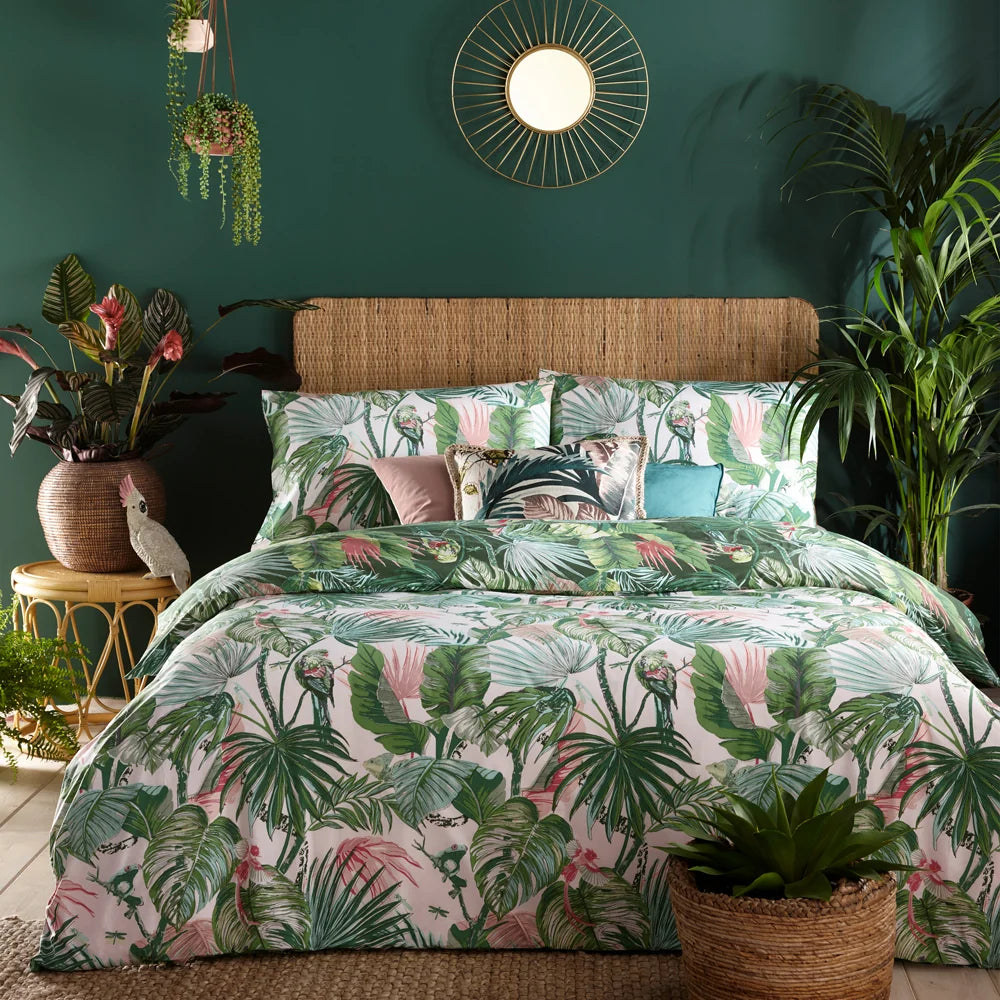 Bed dressed in the jade rainforest print duvet cover set with matching pillowcases against a dark green wall, styled with a gold sunburst mirror, a rattan headboard, hanging plants, a wicker vase of pink flowers and scatter cushions in pink, teal and leaf print – Kumperi