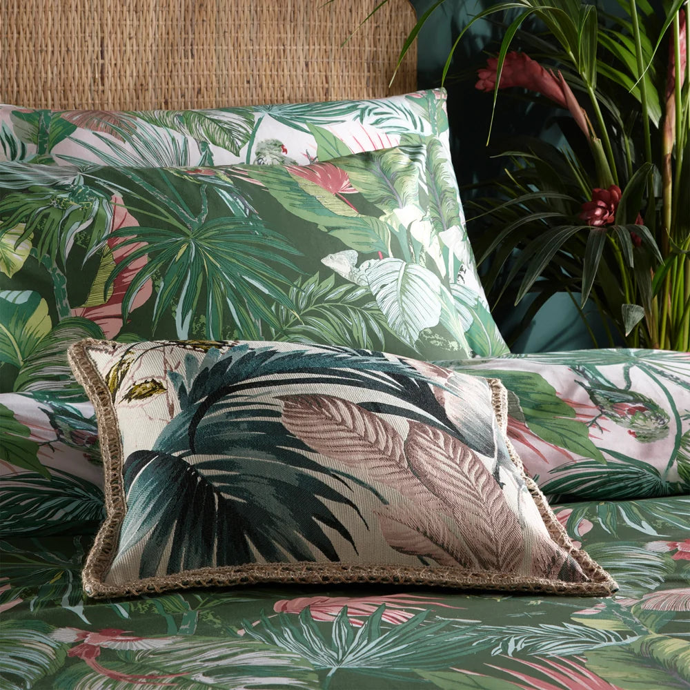 Close-up of the jade and blush reversible tropical print pillowcases stacked behind a cream leaf print cushion with a woven trim, set against a rattan headboard with red ginger flowers nearby – Kumperi
