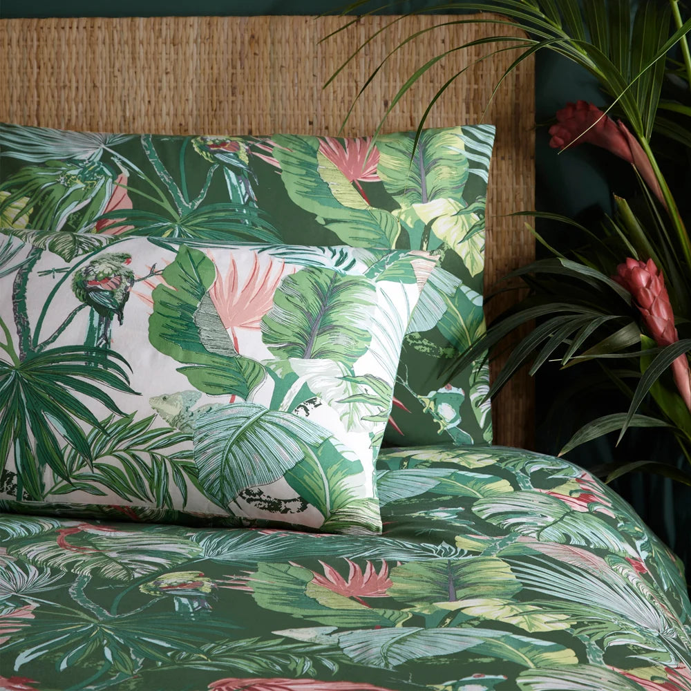 Close-up showing both reversible sides of the pillowcases side by side, one in blush tones with a parakeet and lizard print and the other in dense jade green tropical foliage, against a rattan headboard with red ginger flowers – Kumperi