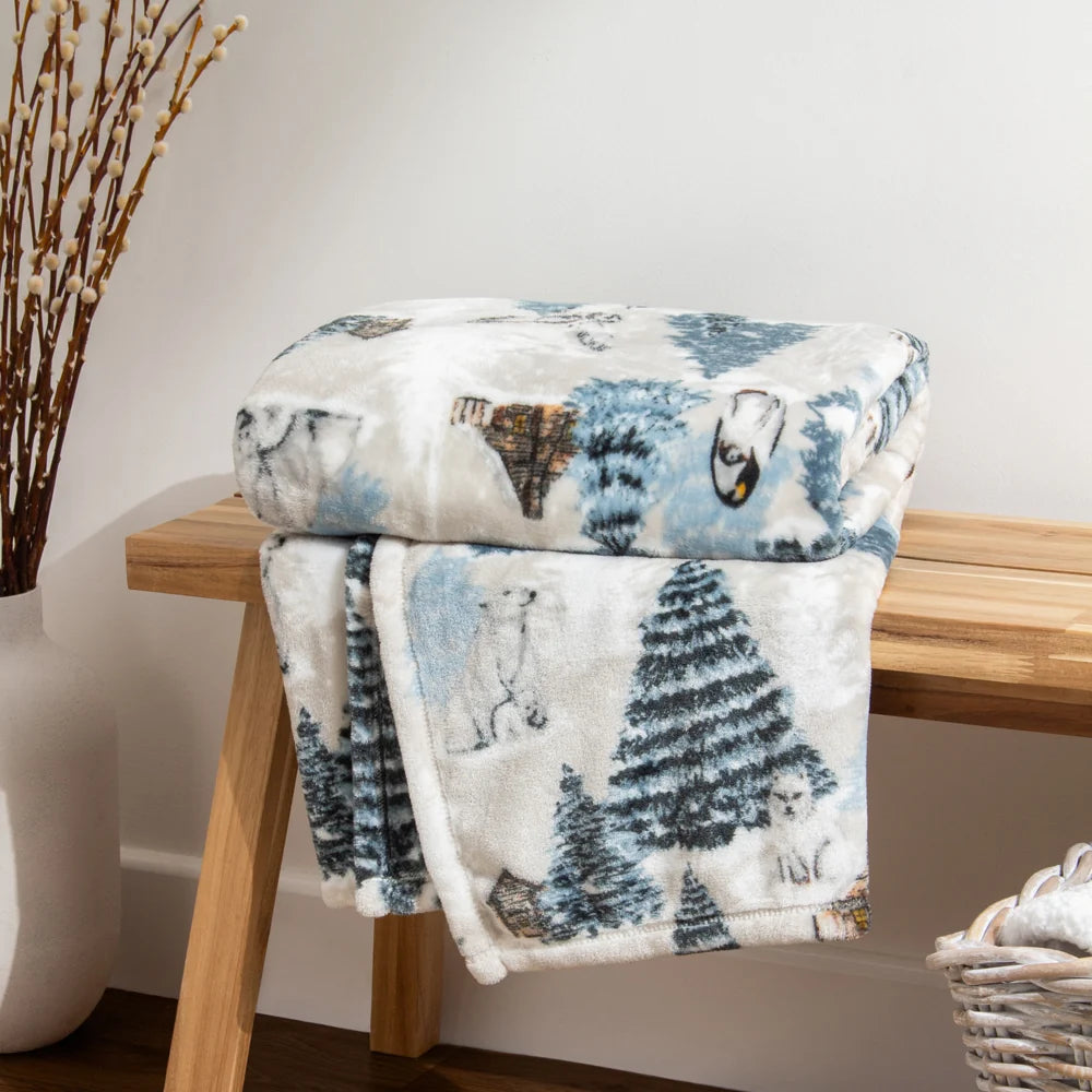 Throw draped and partly folded over a wooden bench in a room with white walls, showing the winter print with pine trees, a polar bear, an owl and cabin motifs, styled beside a vase of dried branches and a wicker basket – Kumperi
