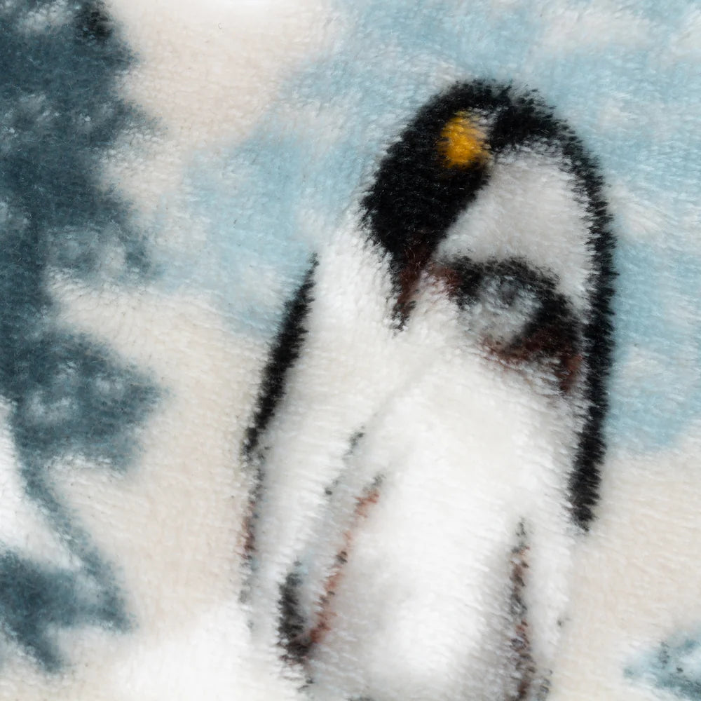Close-up detail of the fleece fabric showing a printed penguin with a golden yellow head patch standing against a pale blue sky and white snow background – Kumperi