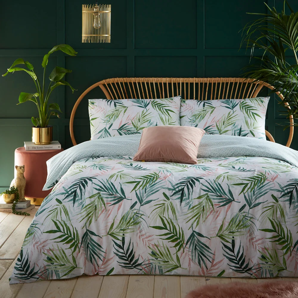 Bed dressed in the white and green watercolour palm print duvet cover with matching pillowcases and the sage geometric reverse folded back, styled with a blush velvet cushion, a rattan headboard against a dark green panelled wall, and potted palm plants either side – Kumperi