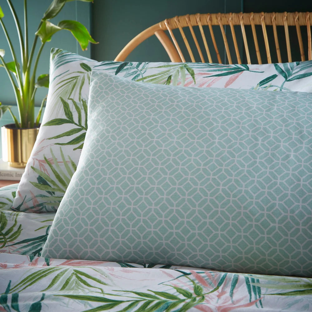 Close-up of a sage green geometric hexagon print pillowcase layered against a white palm leaf print pillowcase, set on the bed near a rattan headboard – Kumperi