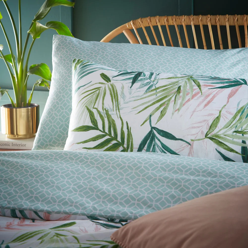 Close-up showing both reversible sides together, the sage green geometric hexagon print pillowcase beside a white cushion with a green and pink palm leaf print, resting on the geometric print duvet cover – Kumperi