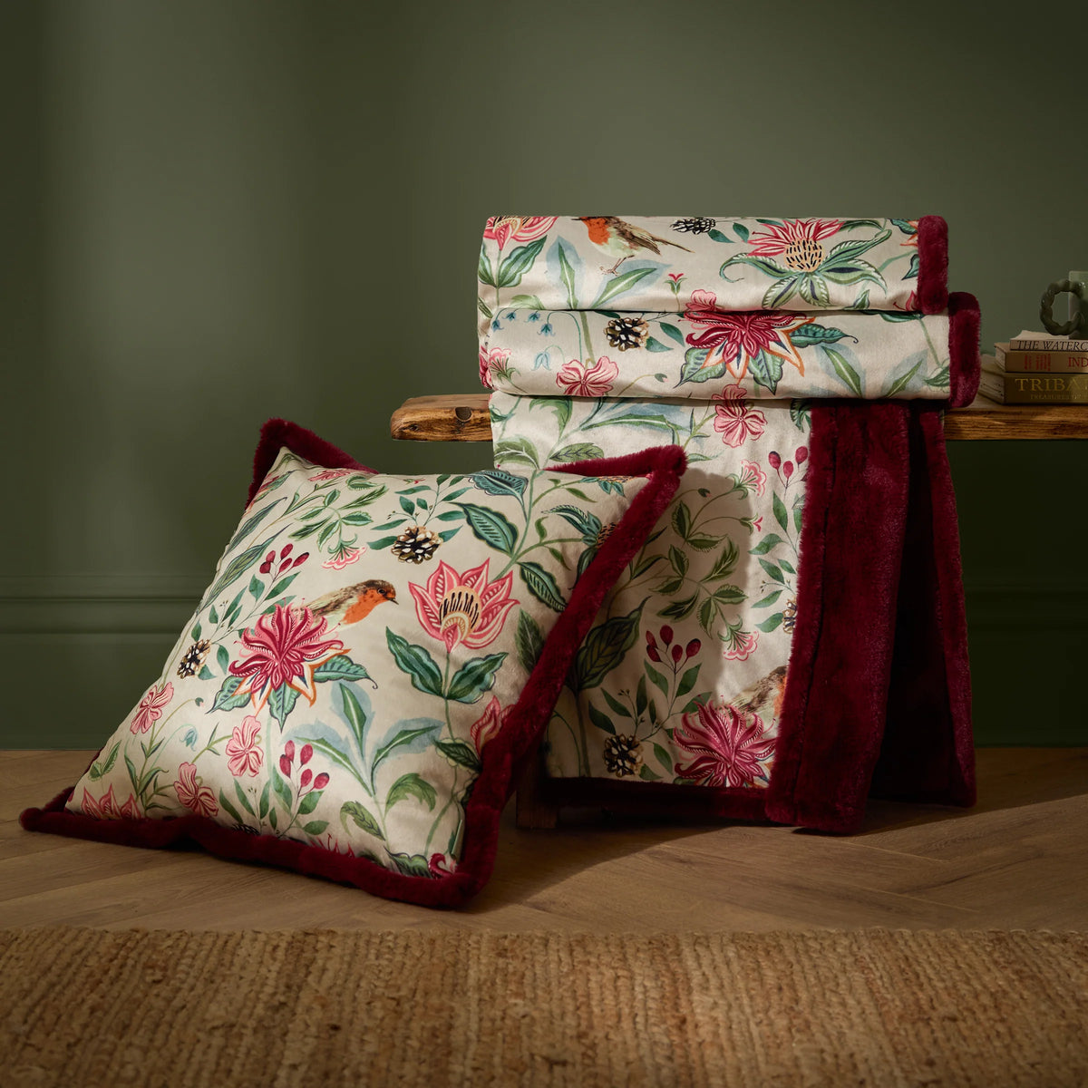 Floral velvet throw draped over a wooden bench beside a matching square cushion with faux fur trim, both featuring the pink honeysuckle and robin print, set against a dark green wall with stacked books nearby – Kumperi