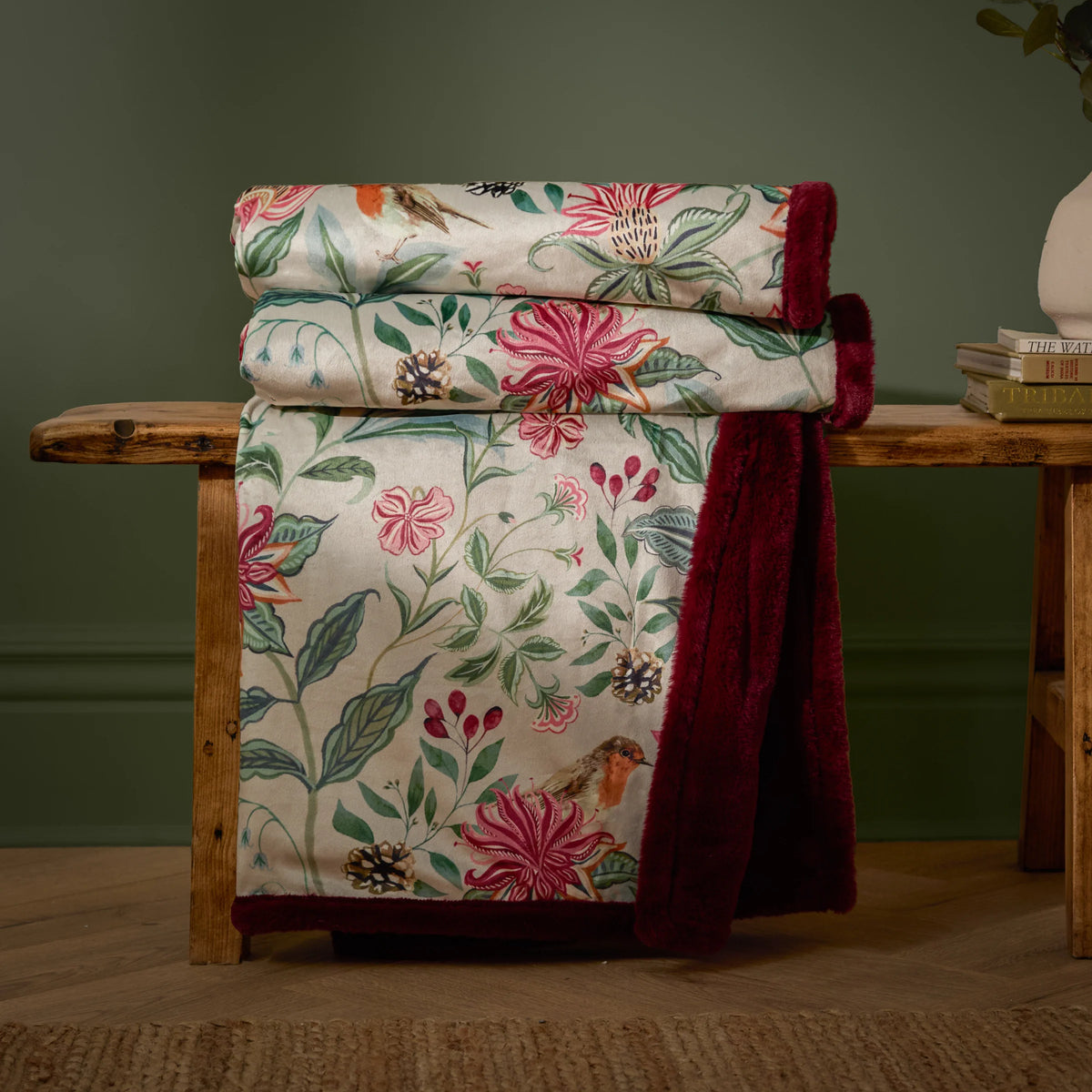 Floral velvet throw rolled and draped over a wooden bench against a dark green wall, showing the cream floral print with pink blooms, pinecones and a robin, with the burgundy faux fur reverse hanging down – Kumperi