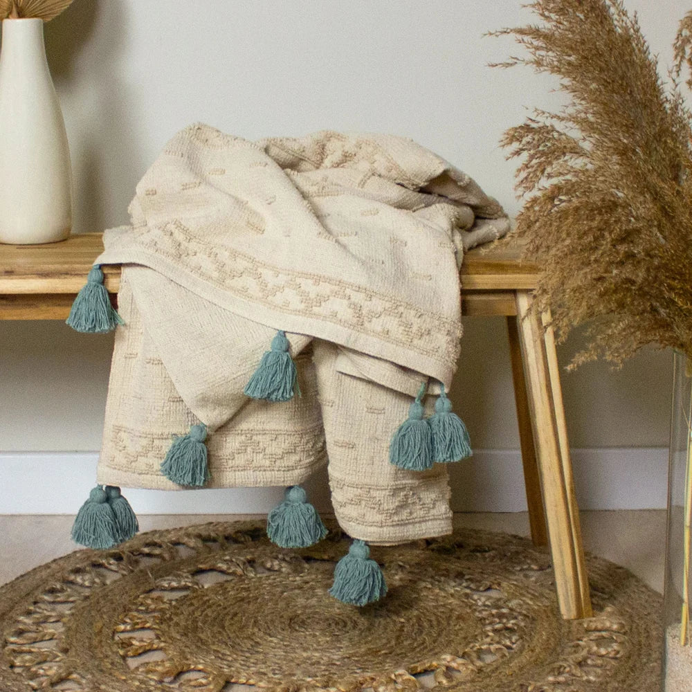 Cream tufted throw draped over a wooden bench with seafoam green tassels hanging down over a round jute rug, styled beside a cream vase and dried pampas grass against a pale green wall – Kumperi