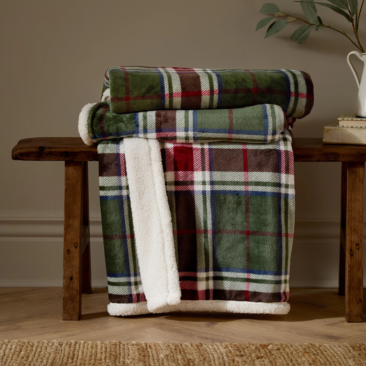 Two tartan check throws rolled and stacked on a wooden bench, with a third draped down showing the pine and berry check print alongside the white sherpa fleece reverse, styled beside a jug of eucalyptus and books against a taupe wall – Kumperi