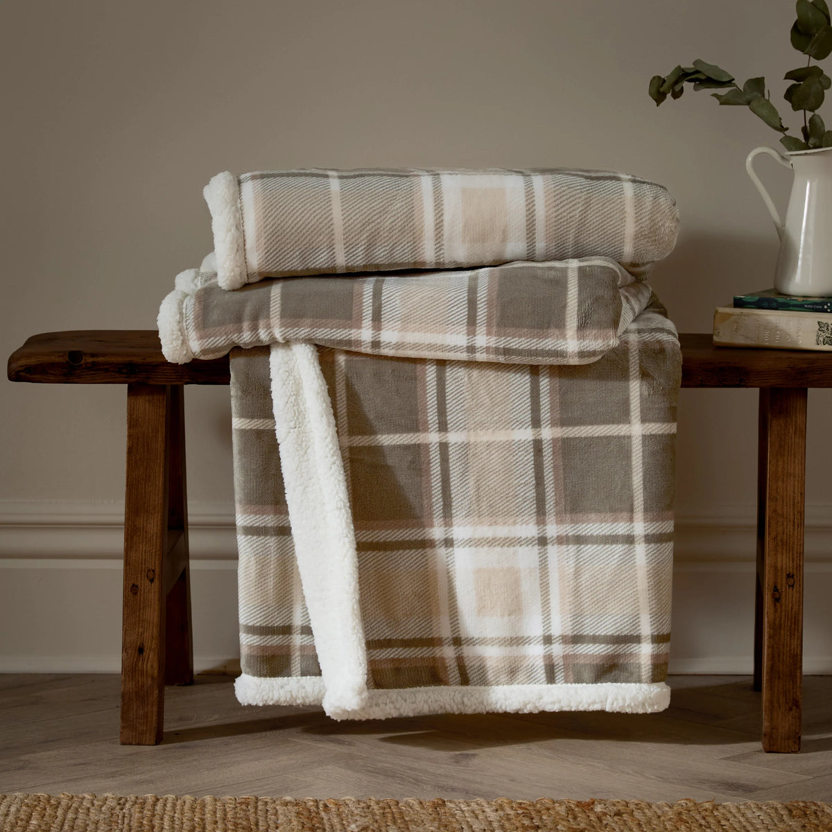 Green and natural tartan check throws rolled and stacked on a wooden bench, with a third draped down showing the check print alongside the white sherpa fleece reverse, styled beside a jug of eucalyptus and books against a taupe wall – Kumperi