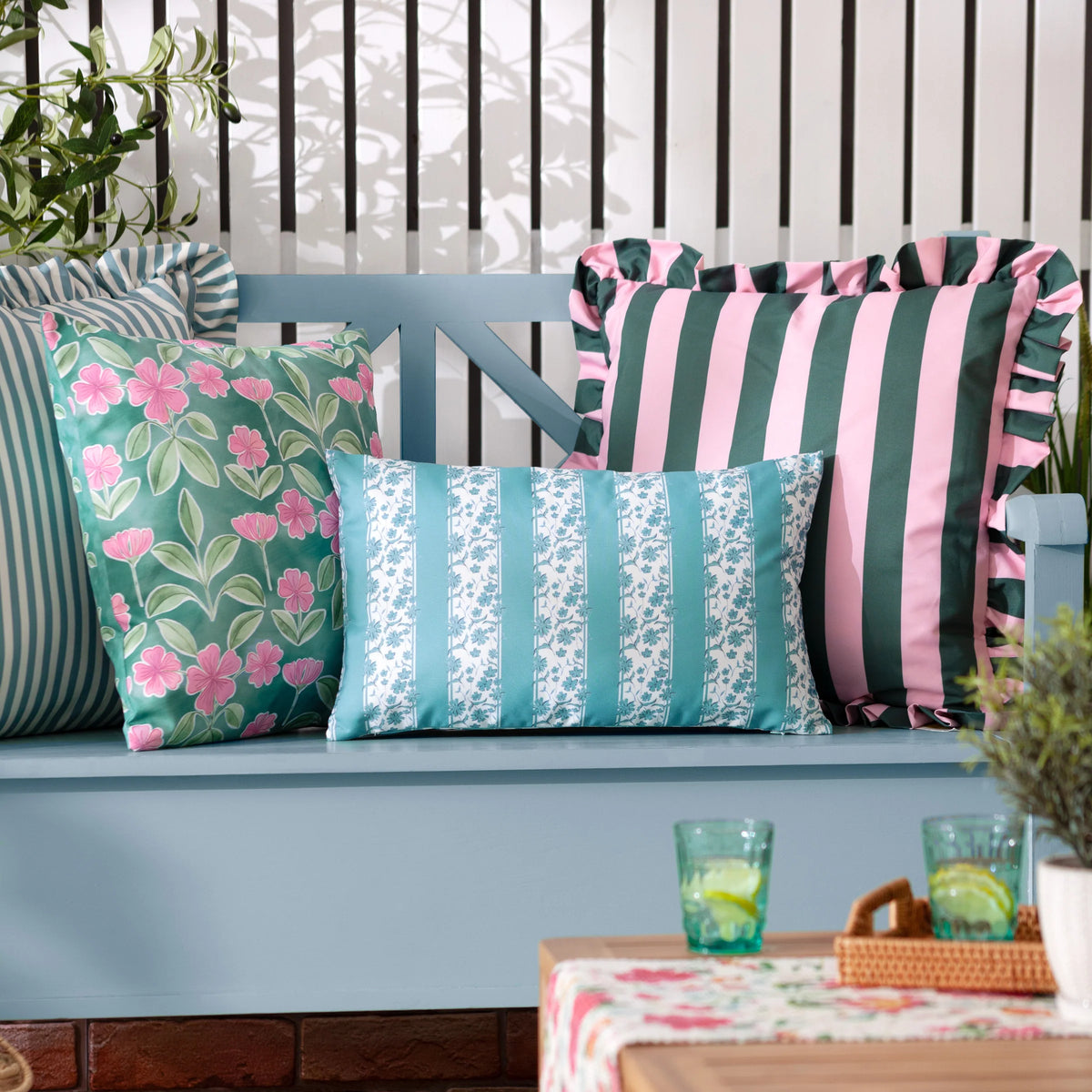 Mineral blue floral stripe rectangular cushion styled on a blue-grey garden bench alongside a green floral square cushion, a pink and green striped ruffle cushion, and a striped bolster, with plants and drinks on a table in the foreground – Kumperi