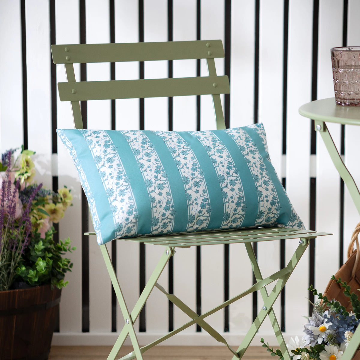 Rectangular mineral blue and white floral stripe outdoor cushion resting on a sage green folding metal chair, with a purple side table, glass tumbler and potted flowers nearby – Kumperi