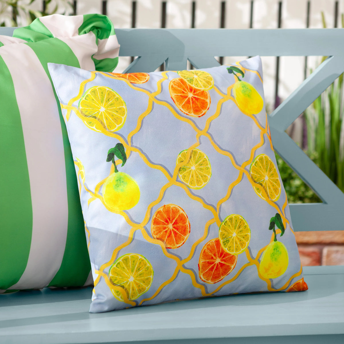 Angled view of the citrus trellis cushion resting against a green and white striped ruffled cushion on a light blue garden bench with greenery in the background – Kumperi