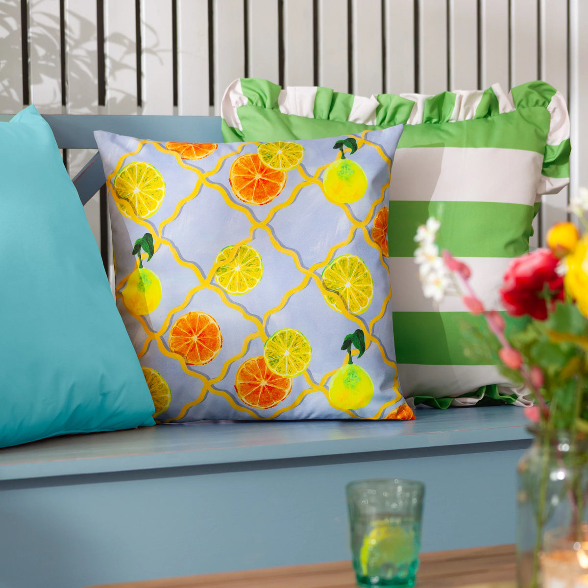 Citrus trellis cushion styled on a blue garden bench beside a teal cushion and a green and white striped ruffled cushion, with fresh flowers in the foreground – Kumperi