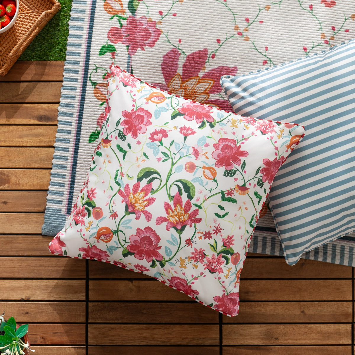 Floral candy red cushion styled diagonally on wooden decking beside a blue and white striped cushion, with a woven picnic basket, potted plant and a striped picnic rug with matching floral border in the background – Kumperi