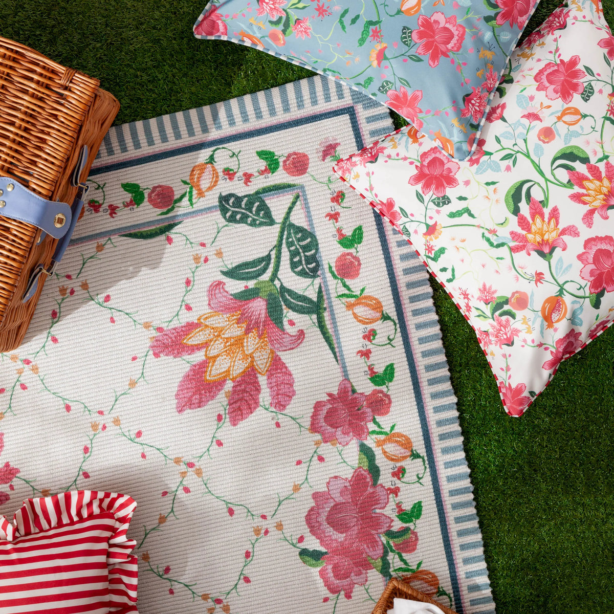 Overhead view on artificial grass showing the floral cushion alongside a matching light blue floral cushion, a striped picnic rug with floral border, a wicker picnic basket and a red and white striped ruffled fabric – Kumperi