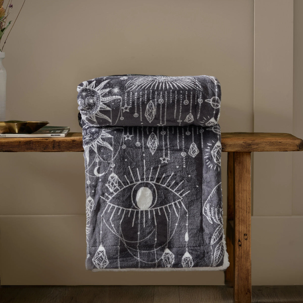 Charcoal constellation throw rolled and draped over a wooden bench, showing the eye and sun print, styled beside a vase of dried flowers and a stack of magazines on a beige wall background – Kumperi