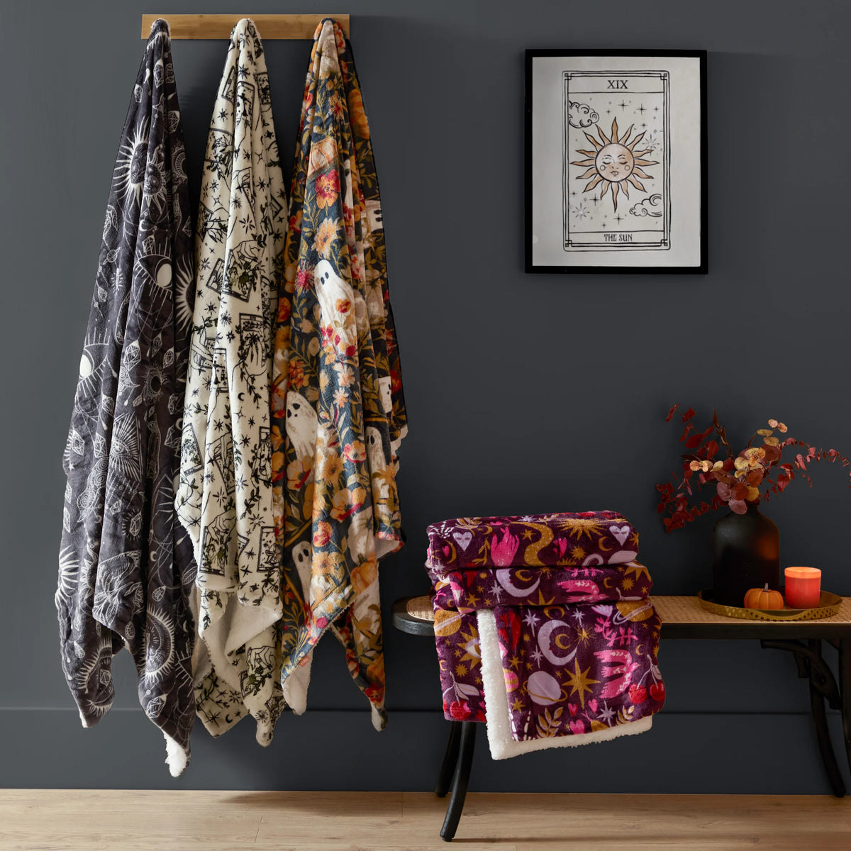 Charcoal constellation throw hanging on a wall hook beside a cream tarot-print throw and a navy floral ghost-print throw, with a folded burgundy celestial throw stacked on a stool nearby and a tarot card art print on the wall – Kumperi