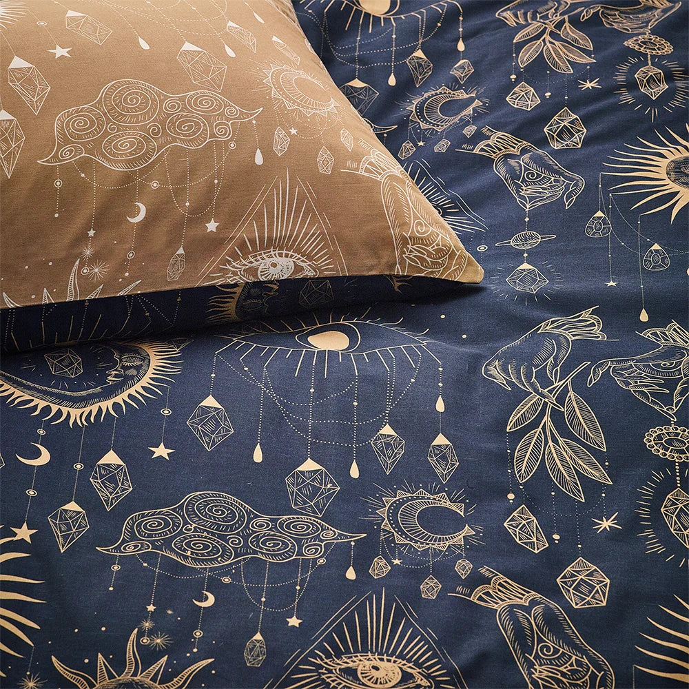 Close-up flat view of a gold cushion with cream celestial line art of clouds, an eye and hanging crystals, resting beside the navy duvet cover showing suns, moons, hands and crystal chain motifs in gold – Kumperi
