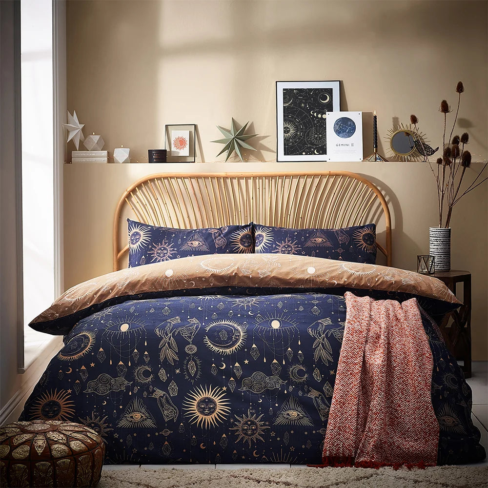 Bed dressed in the gold and navy celestial duvet cover set with matching pillowcases showing suns, moons, eyes, hands and hanging crystal motifs, styled with a rust chevron throw, a rattan headboard and celestial art prints on a shelf above against a beige wall – Kumperi