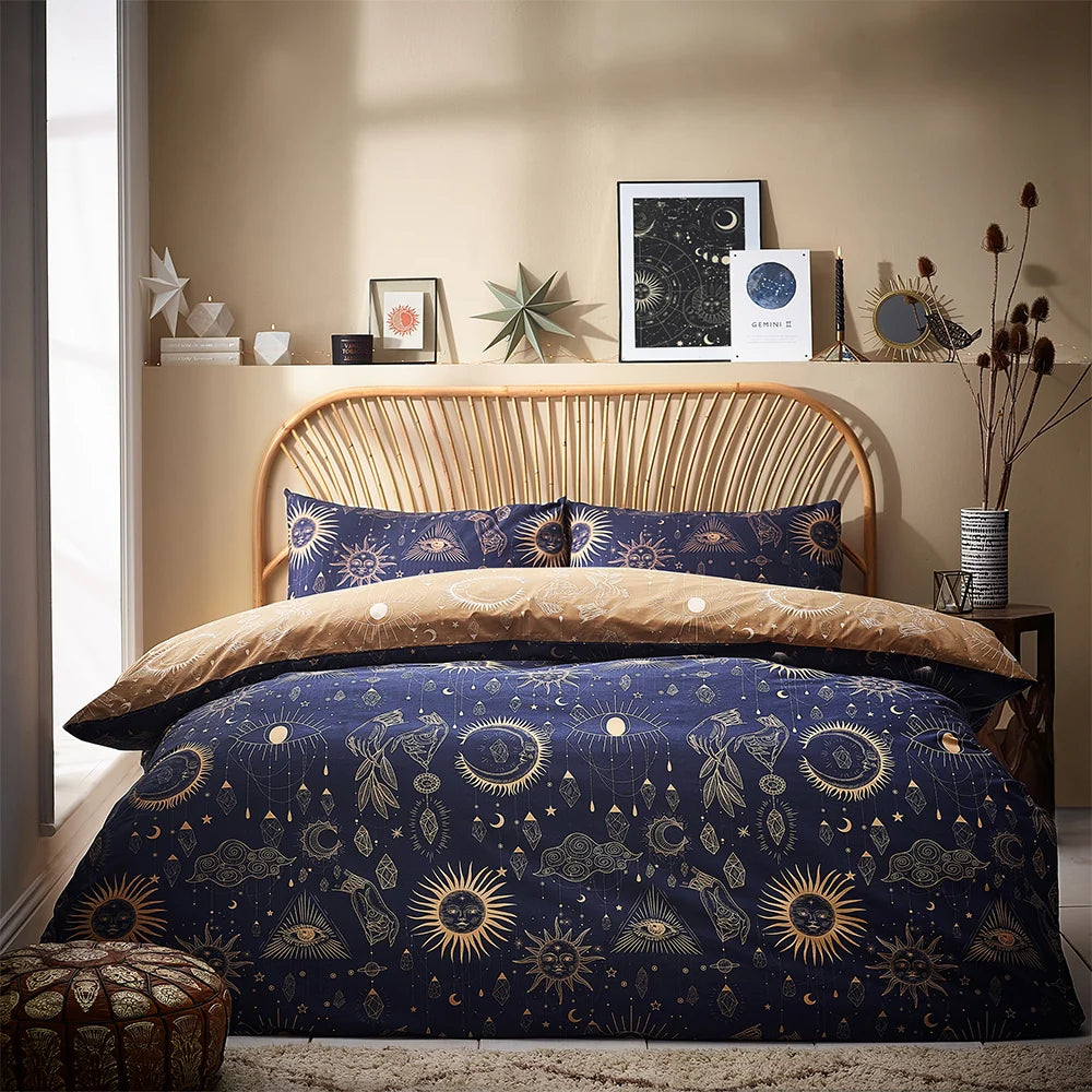 Close view of the bed against a rattan headboard showing gold pillowcases with a navy celestial print of suns, eyes and hands, layered over the navy and gold duvet, styled beside a patterned vase of dried branches – Kumperi