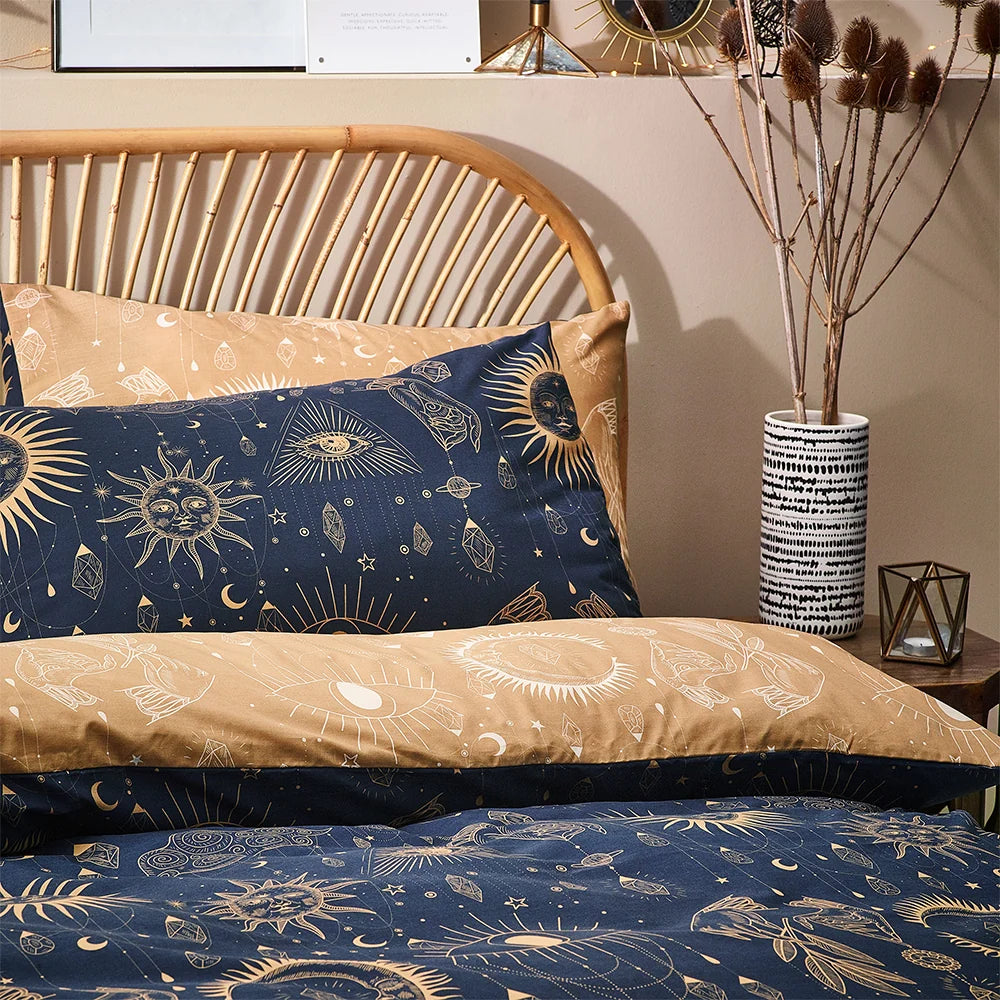 Detail view of gold and navy celestial pillowcases propped against a rattan headboard, showing suns with faces, an all-seeing eye, hands and hanging crystal chain motifs, with a patterned vase and dried branches on a bedside table – Kumperi
