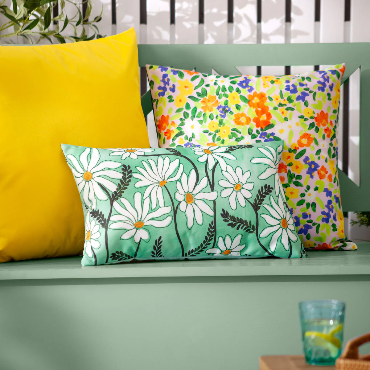 Daisy Fields cushion styled on a green painted bench alongside a yellow cushion and a multicoloured ditsy floral cushion, with a glass of water and greenery nearby – Kumperi