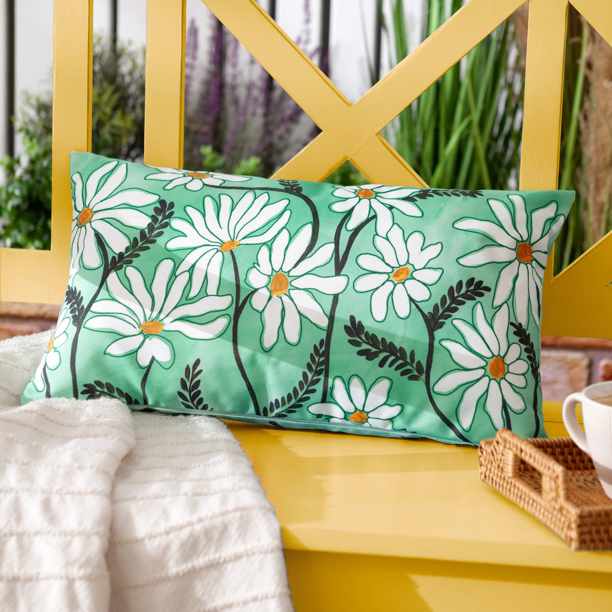 Daisy Fields cushion resting on a yellow garden bench beside a cream throw and a woven basket, with purple and green foliage in the background – Kumperi