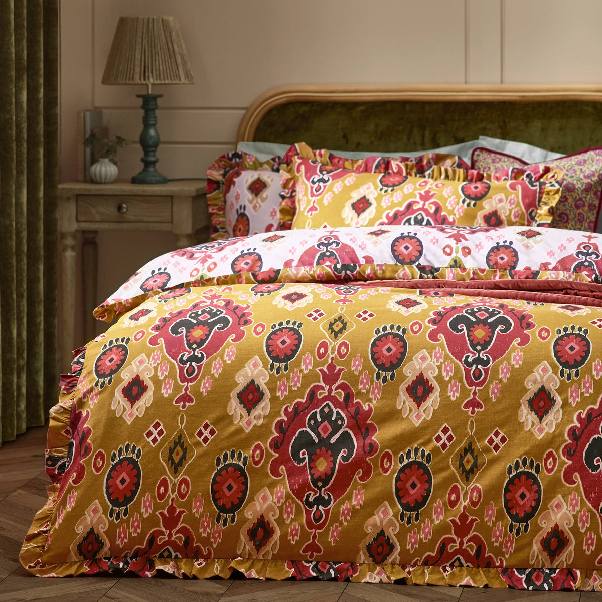 Angled view of the bed showing the ruffled amber Ikat duvet cover, layered pillowcases in amber and white reversible prints, and a floral cushion, set against a green velvet headboard with a bedside lamp – Kumperi