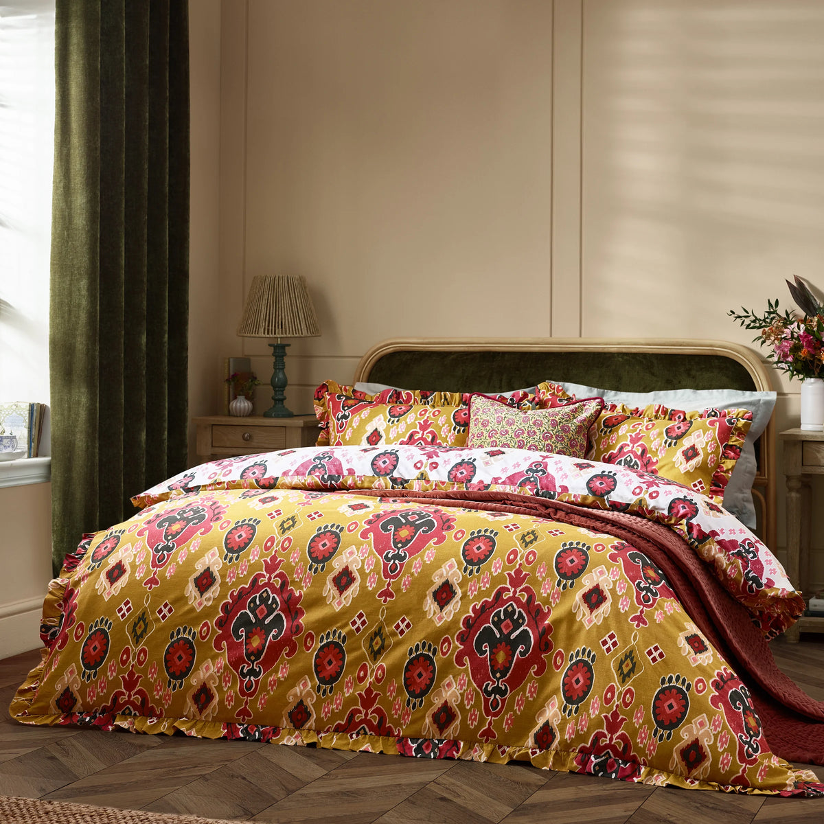 Bed dressed in the amber Ikat print duvet cover set with a ruffled edge, matching amber pillowcases, a white reversible pillowcase and a smaller floral cushion, styled with a rust waffle throw against a green velvet headboard and dark green curtains – Kumperi