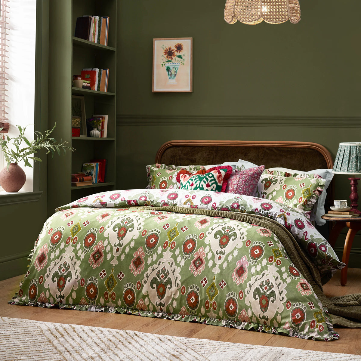 Angled view of the bed dressed in the cedar green ruffled Ikat duvet cover set with a reversible cream pillowcase and layered pink and green scatter cushions, set against a dark green wall with a bedside lamp and rattan pendant light – Kumperi