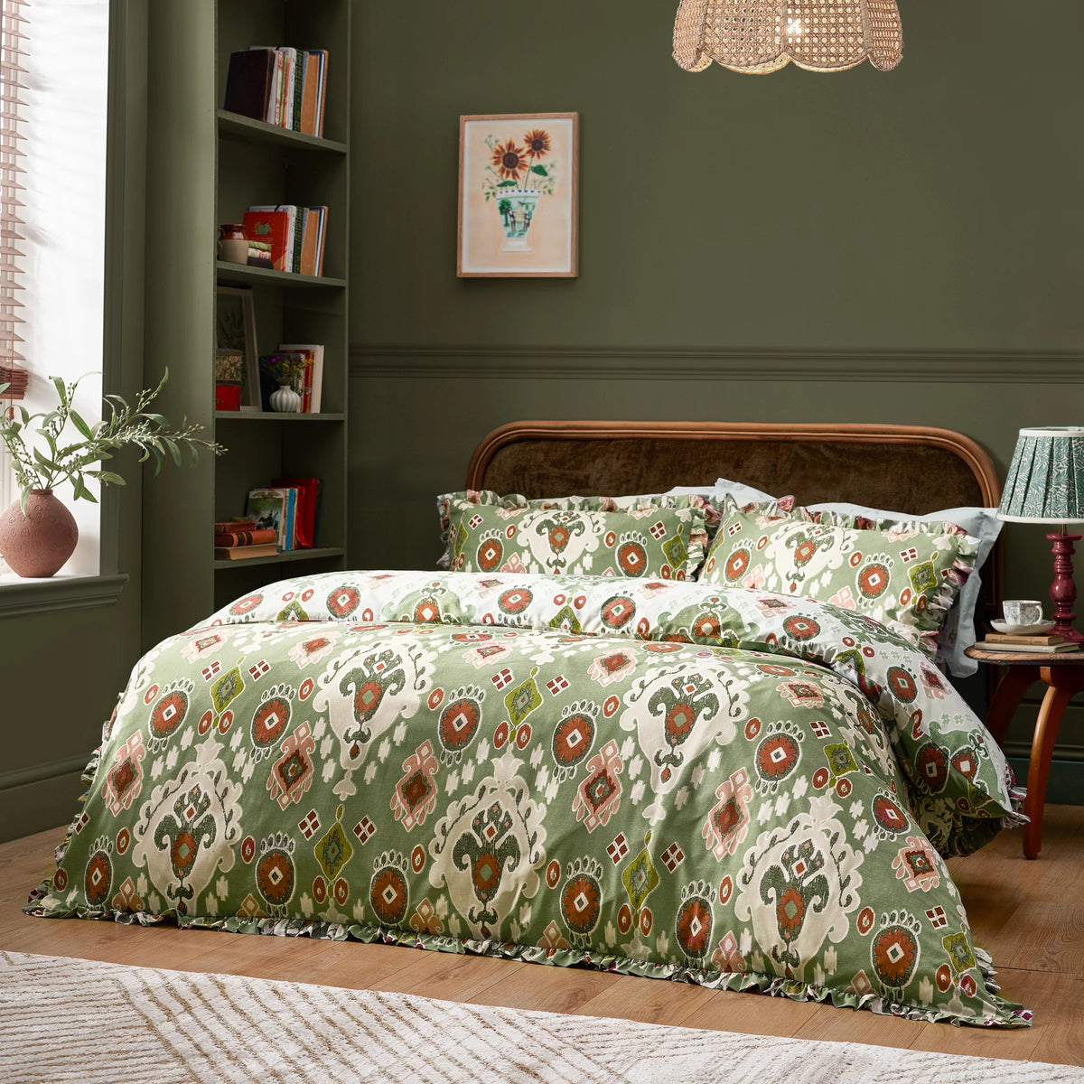 Full bed dressed in the cedar green Ikat print duvet cover set with ruffled trim and matching pillowcases, styled against a dark green wall with a rattan headboard, a hanging rattan pendant light and a framed sunflower print above a bookshelf – Kumperi