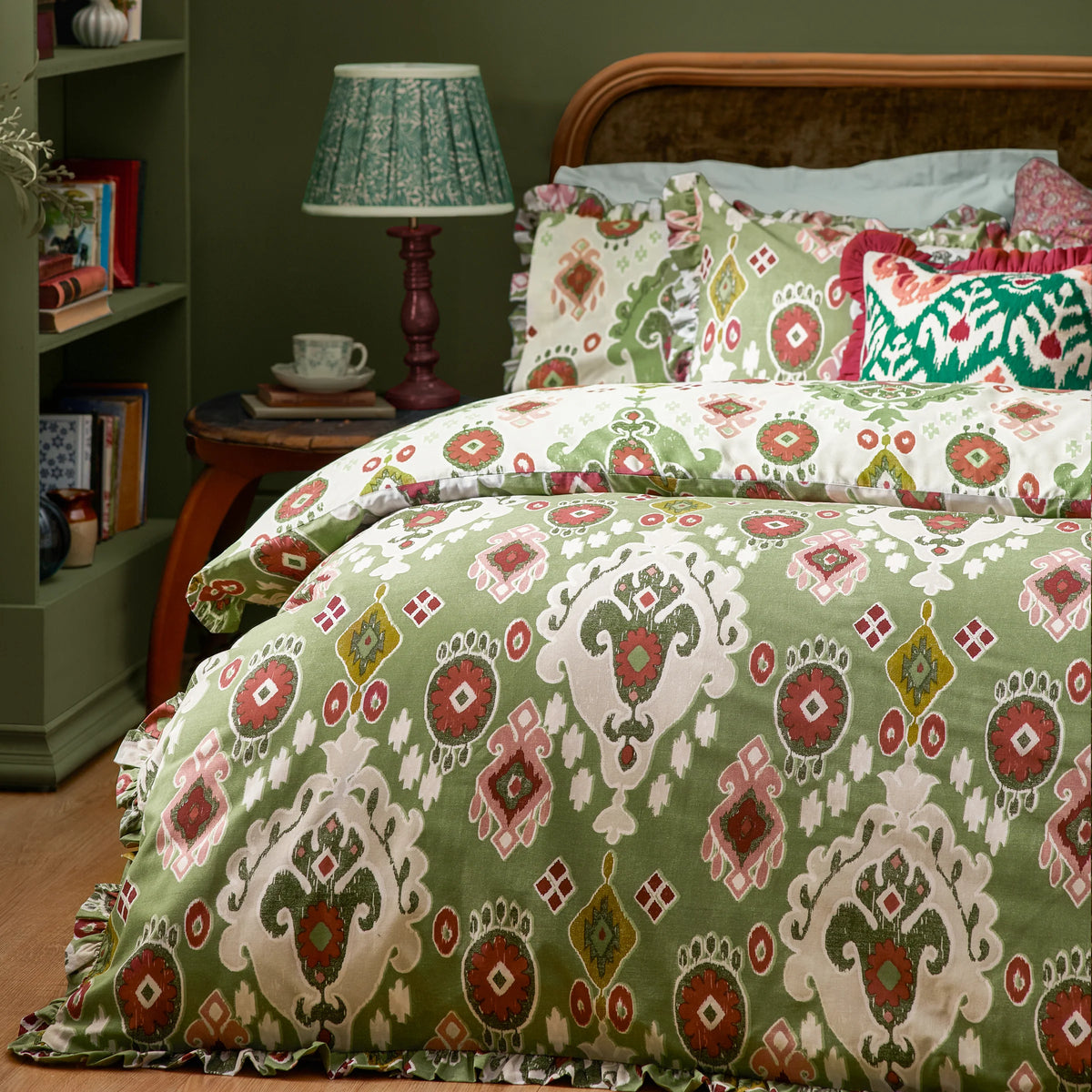 Bed dressed in the cedar green Ikat print duvet cover set with a ruffled edge, matching pillowcases, a cream reversible pillowcase and a green tribal print cushion, styled against a dark green wall with a rattan headboard, a bedside lamp with a patterned shade, a teacup and books – Kumperi