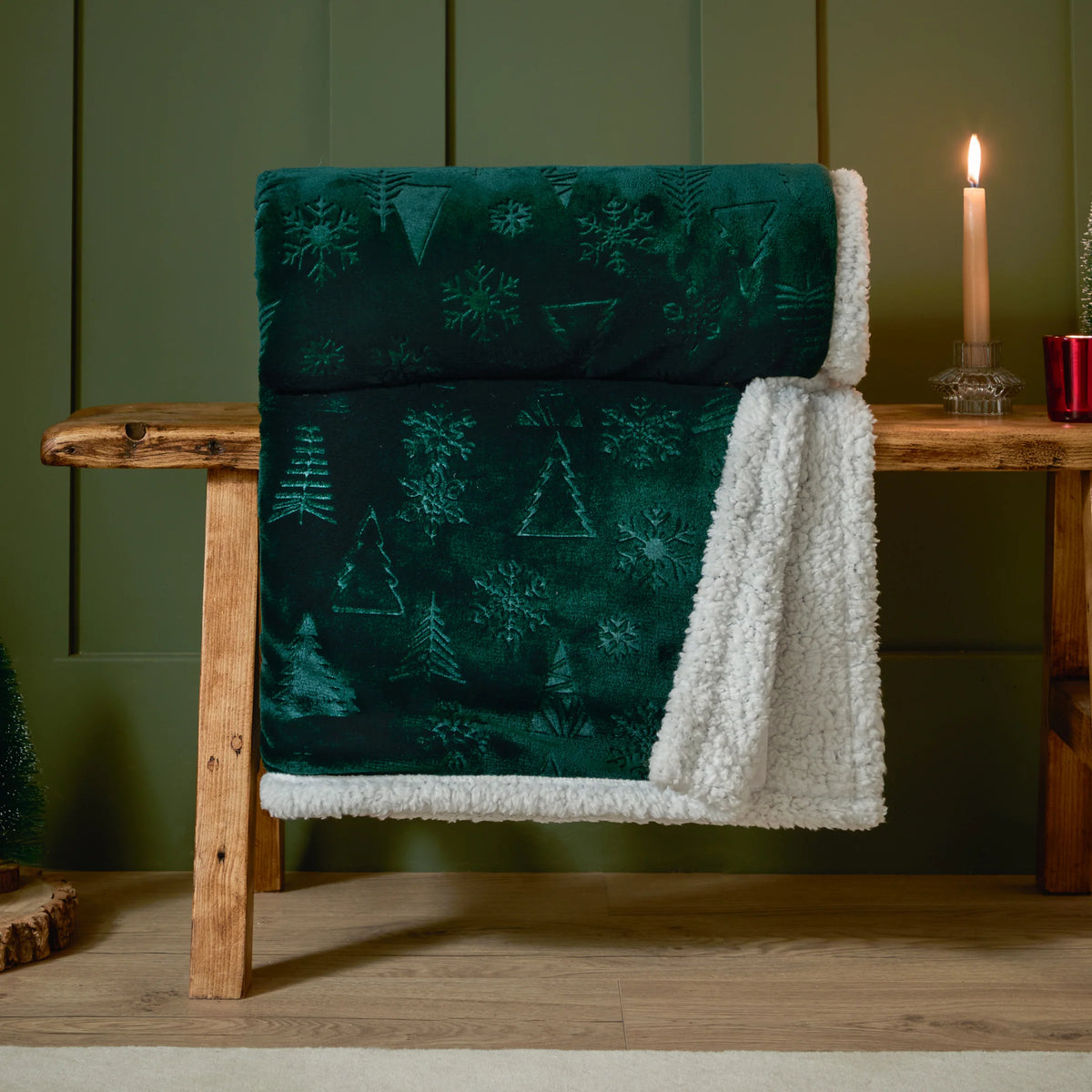 Emerald embossed throw rolled and draped over a wooden bench against an olive green panelled wall, with the white sherpa fleece reverse showing, styled beside a lit candle, a red votive holder and a small Christmas tree – Kumperi