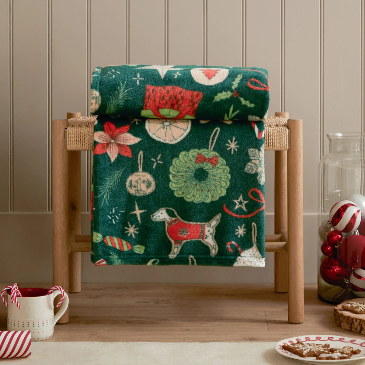 Emerald festive treats throw rolled and draped over a woven wooden bench, showing a wreath, gingerbread box, poinsettia flower and a dog in a red jumper, styled beside a mug of candy canes, a striped ribbon and a plate of Christmas cookies – Kumperi