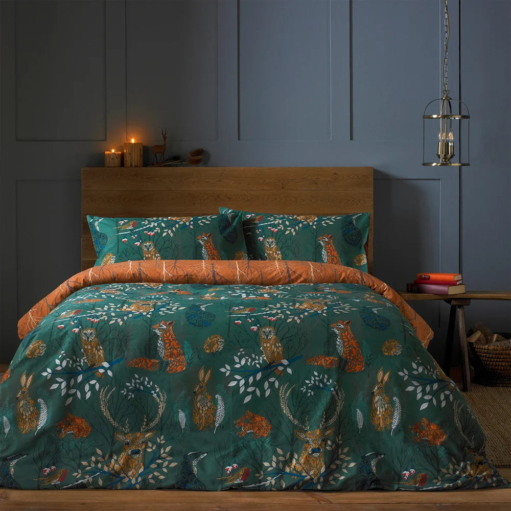 Close view of the bed showing the emerald woodland print duvet cover and pillowcases with foxes, owls and hares, the rust branch print reverse folded back, set against a wooden headboard with a hanging pendant lantern light – Kumperi