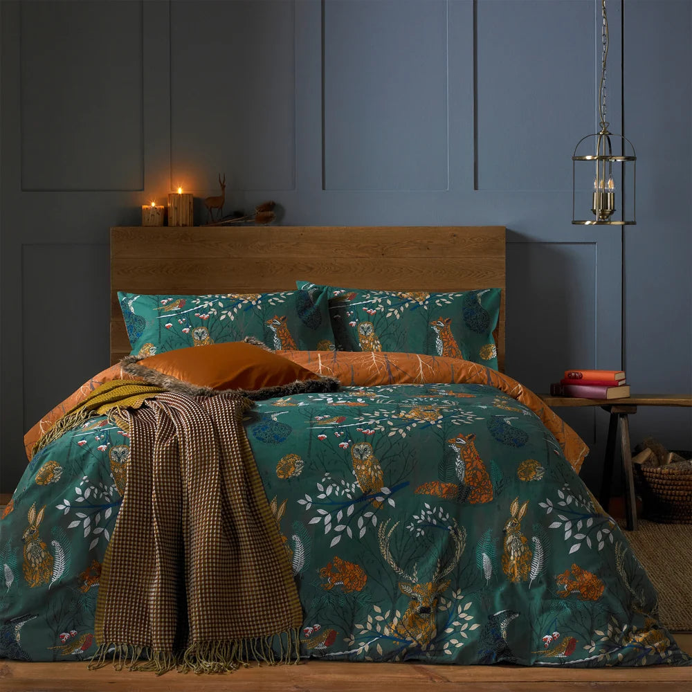 Bed dressed in the emerald woodland animal print duvet cover set with matching pillowcases showing foxes, owls, hares and stags among branches, styled with a burnt orange velvet cushion, a mustard checked throw, a wooden headboard, lit candles and a small deer ornament against a dark blue panelled wall – Kumperi