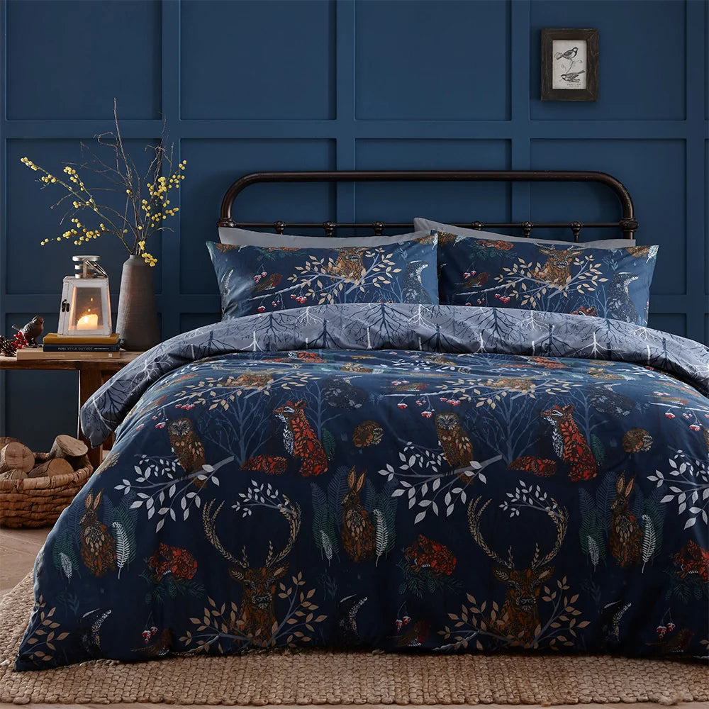 Close view of the navy woodland print duvet cover and pillowcases folded on the bed, showing a fox, owl, hare and stag among branches and berries, with a metal bed frame, a lantern candle and a vase of yellow branches on a bedside table – Kumperi