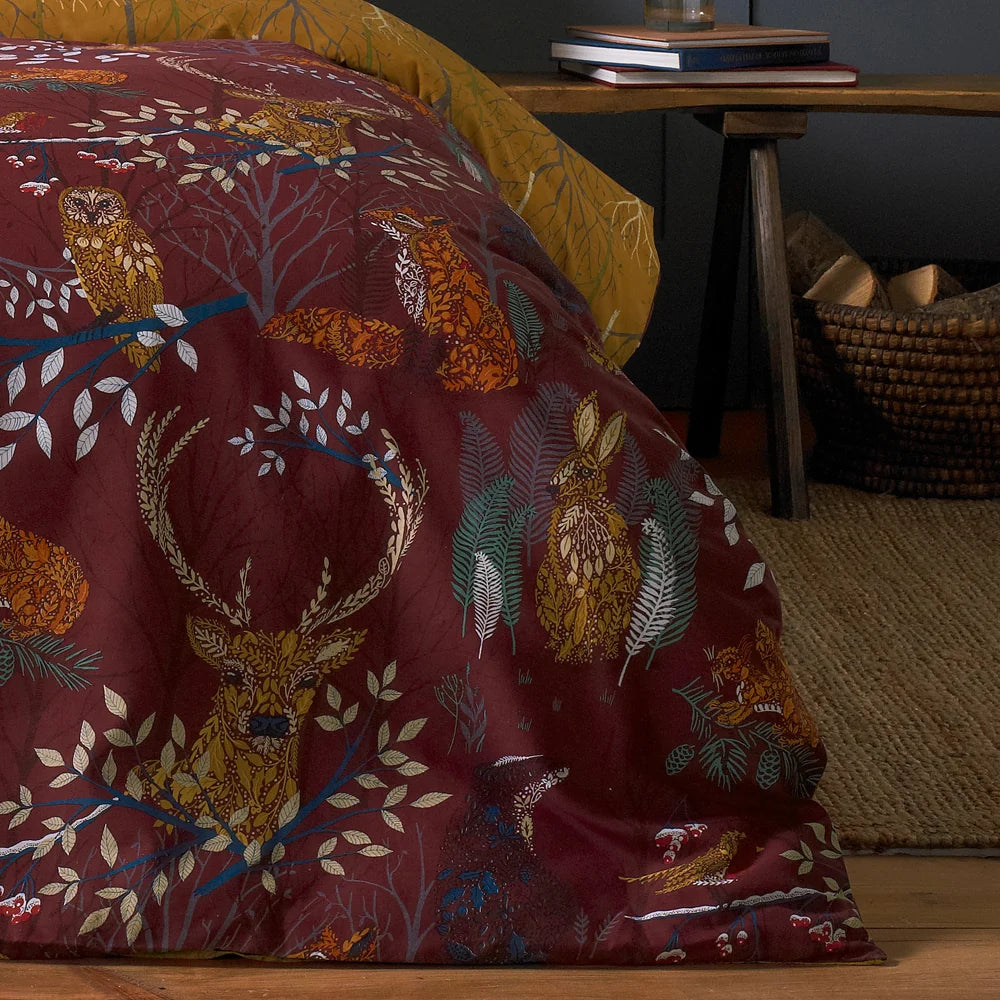 Close-up detail of the rust duvet fabric showing an owl, a fox, a stag, hares and a hedgehog illustrated among leaves and branches, with the mustard reverse visible at the corner – Kumperi
