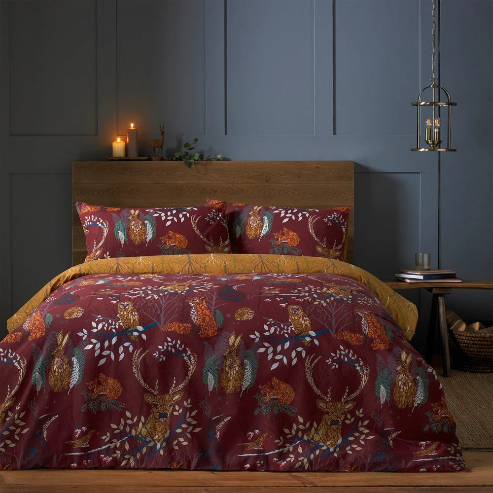 Close view of the bed showing the rust woodland print duvet cover and pillowcases with owls, hares and stags among branches, set against a wooden headboard with lit candles and books on a side table – Kumperi