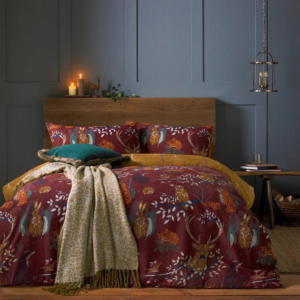 Bed dressed in the rust woodland animal print duvet cover set with matching pillowcases showing foxes, owls, hares and stags among branches, the mustard reverse folded back, styled with a teal velvet cushion, a cream herringbone throw, a wooden headboard and lit candles – Kumperi