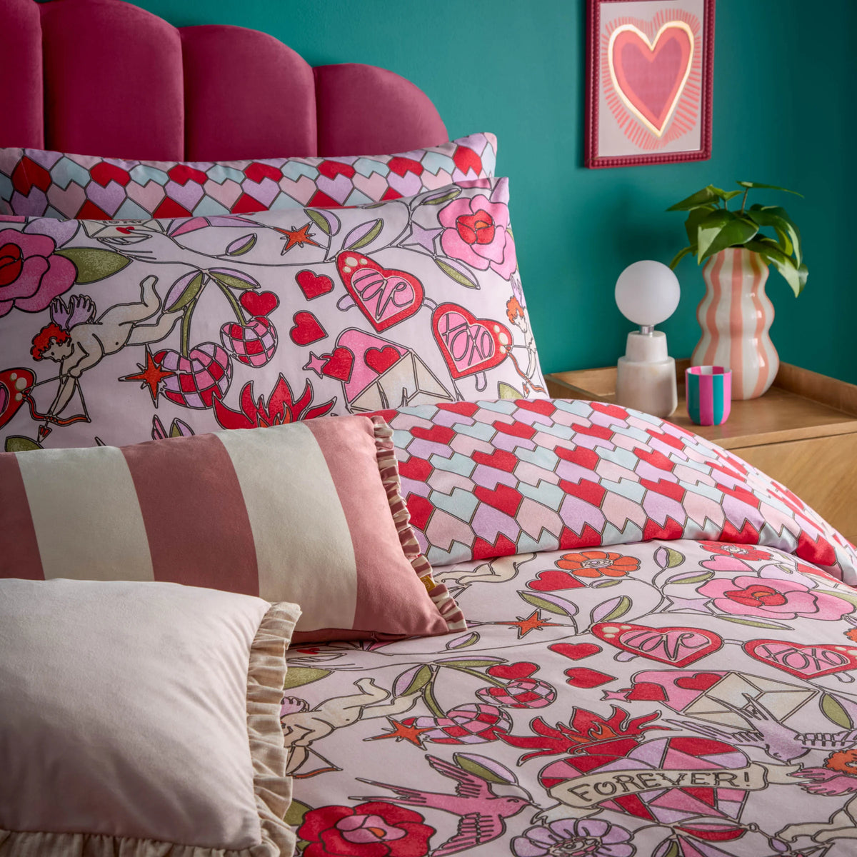 Close-up detail of the pink duvet cover fabric showing hand-drawn cupids, roses, a butterfly heart, love letters and a "Forever" banner, layered with the tessellating heart print pillowcases and a striped ruffle-trimmed cushion – Kumperi