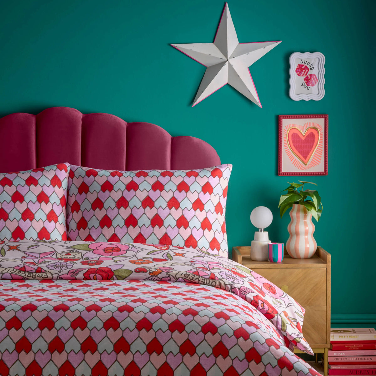 Angled view of the bed showing the pink glass hearts print duvet cover and tessellating heart pillowcases, set against a scalloped magenta velvet headboard and teal wall with a heart artwork above a wooden bedside table – Kumperi