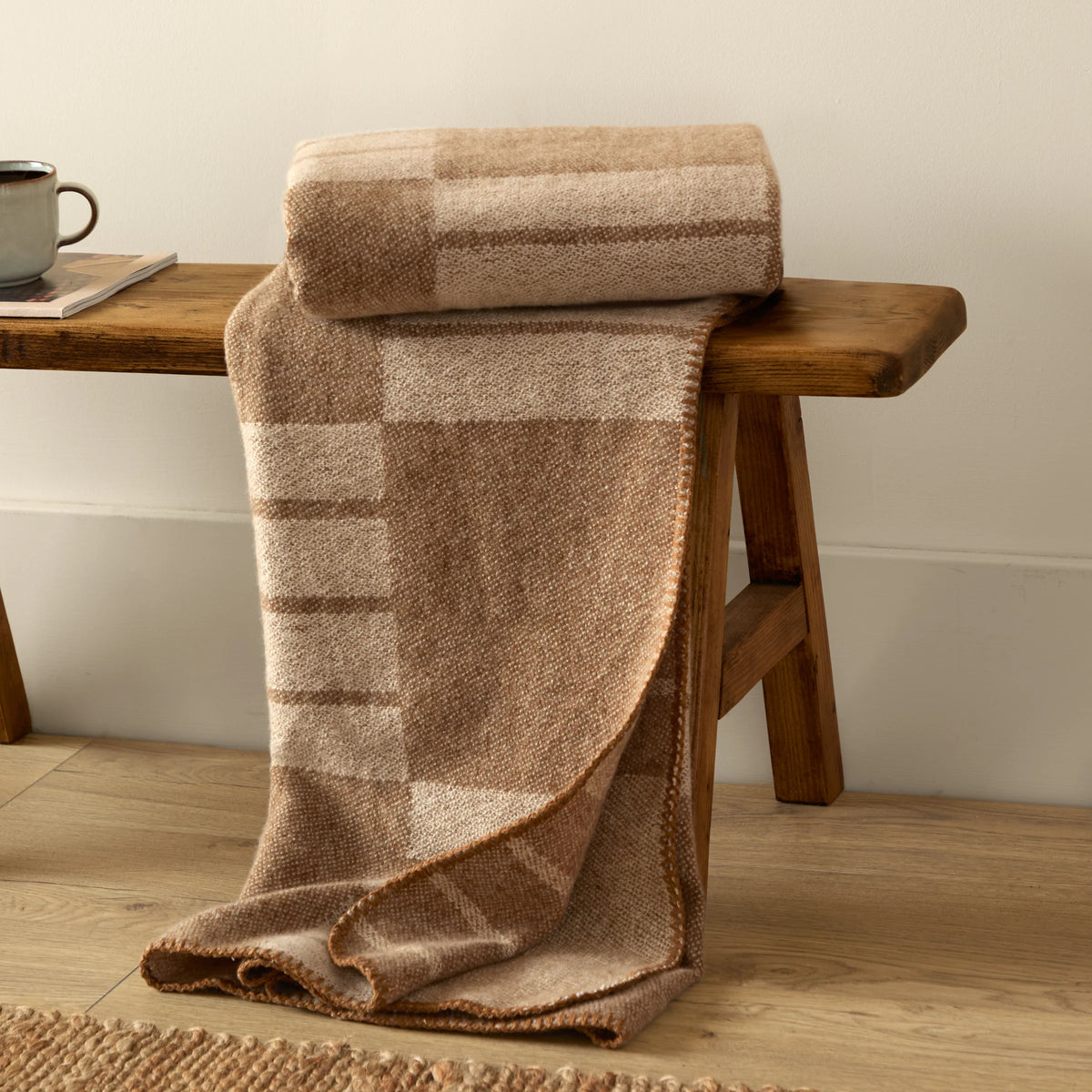 Biscuit woven throw rolled and draped over a wooden bench, showing the geometric block pattern and reverse side, styled beside a mug of coffee and a magazine on a jute rug – Kumperi