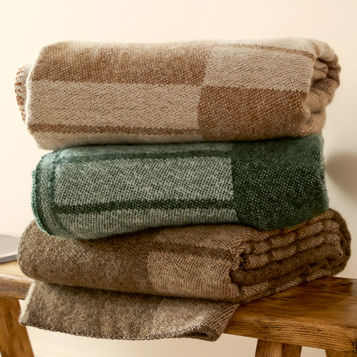 Biscuit woven throw stacked with a matching green geometric throw and a brown patterned throw on a wooden bench, showing the folded edges and blanket stitch trims – Kumperi