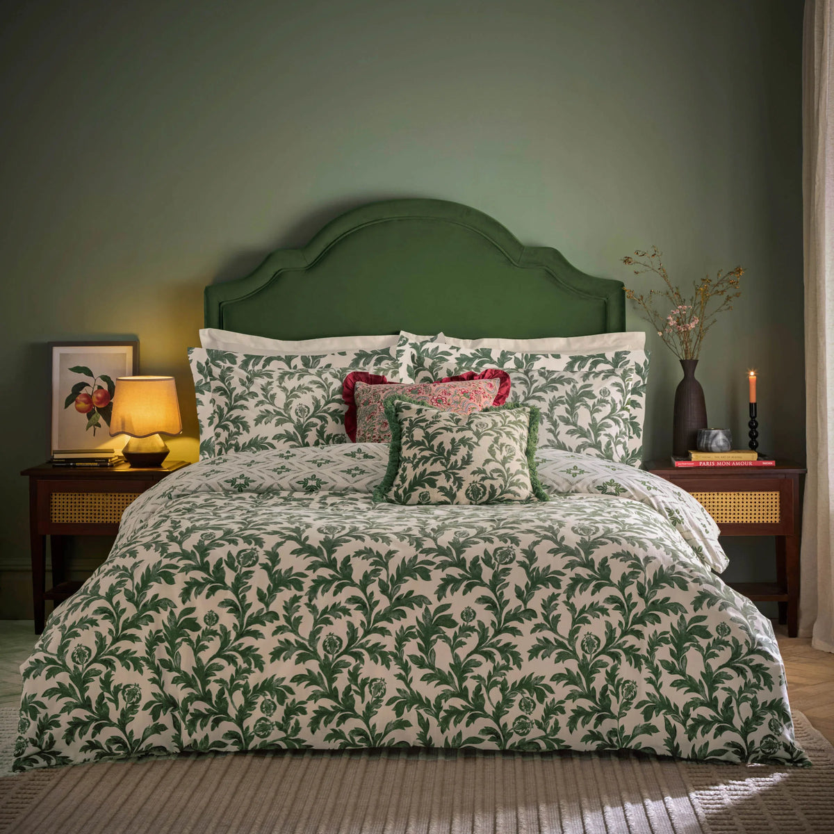Bed dressed in the verdie ivy print duvet cover set with matching Oxford border pillowcases, a diamond geometric reversible pillowcase, a red paisley cushion with ruffle trim, and a green fringed ivy print cushion, styled against a green wall with a green velvet headboard, a botanical framed print, a table lamp and a vase of dried flowers – Kumperi