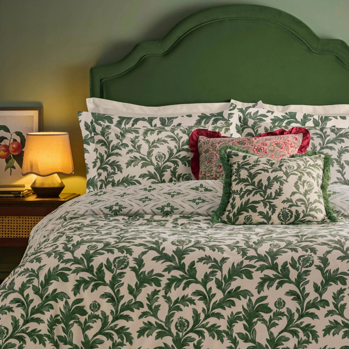 Bed dressed in the verdie ivy print duvet cover set with matching Oxford border pillowcases, a diamond geometric reversible pillowcase, a red paisley cushion with ruffle trim, and a green fringed ivy print cushion, styled against a green wall with a green velvet headboard, a botanical framed print, a table lamp and a vase of dried flowers – Kumperi